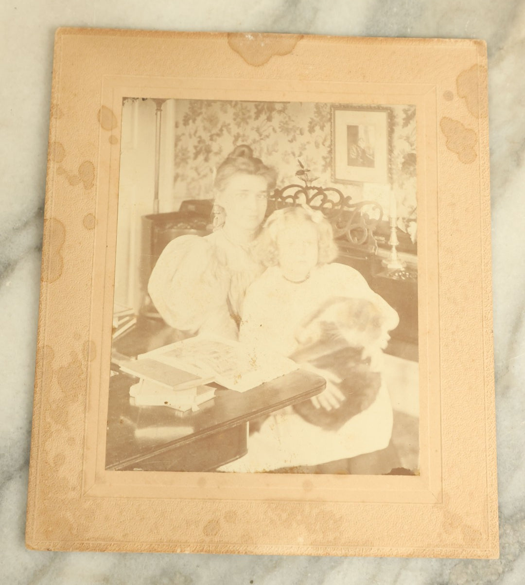 Lot 089 - Antique Boarded Photograph Of A Young Woman And Her Daughter Seated By A Piano Holding A Blurry, Moving Cat