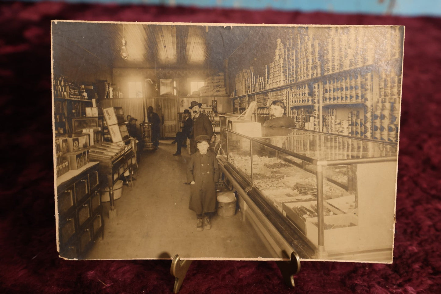 Lot 137 - Small Boarded Photo - General Store Interior Occupational Photo, Many Cans, Ads, Young Boy At Center, Men Around Wood Stove