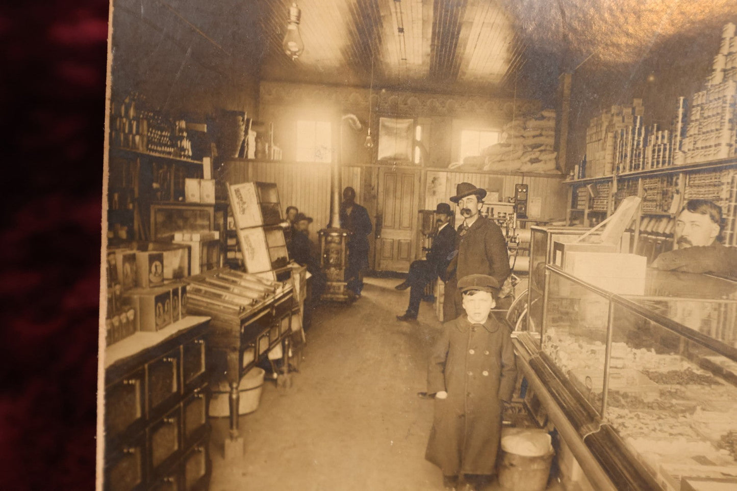 Lot 137 - Small Boarded Photo - General Store Interior Occupational Photo, Many Cans, Ads, Young Boy At Center, Men Around Wood Stove