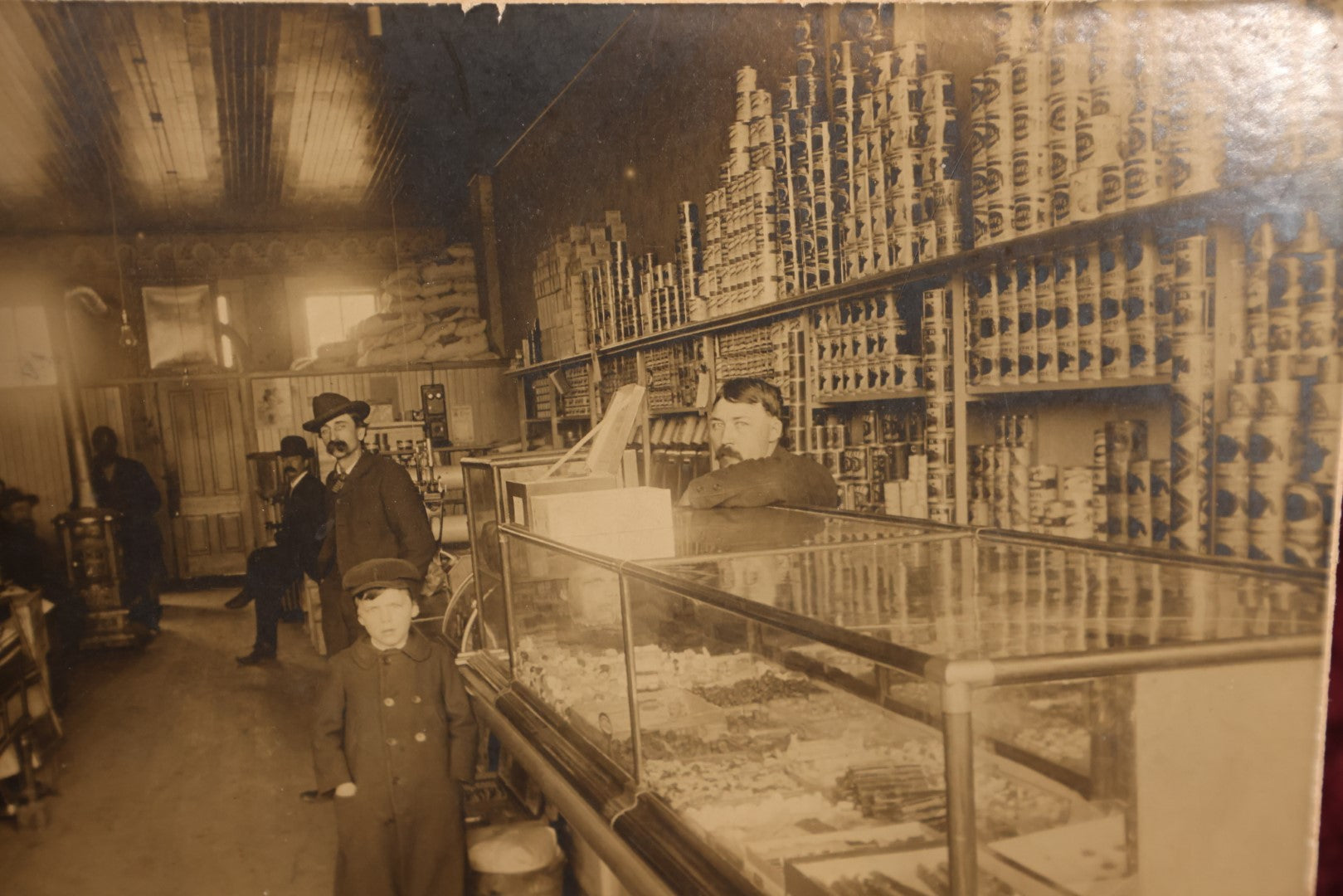 Lot 137 - Small Boarded Photo - General Store Interior Occupational Photo, Many Cans, Ads, Young Boy At Center, Men Around Wood Stove