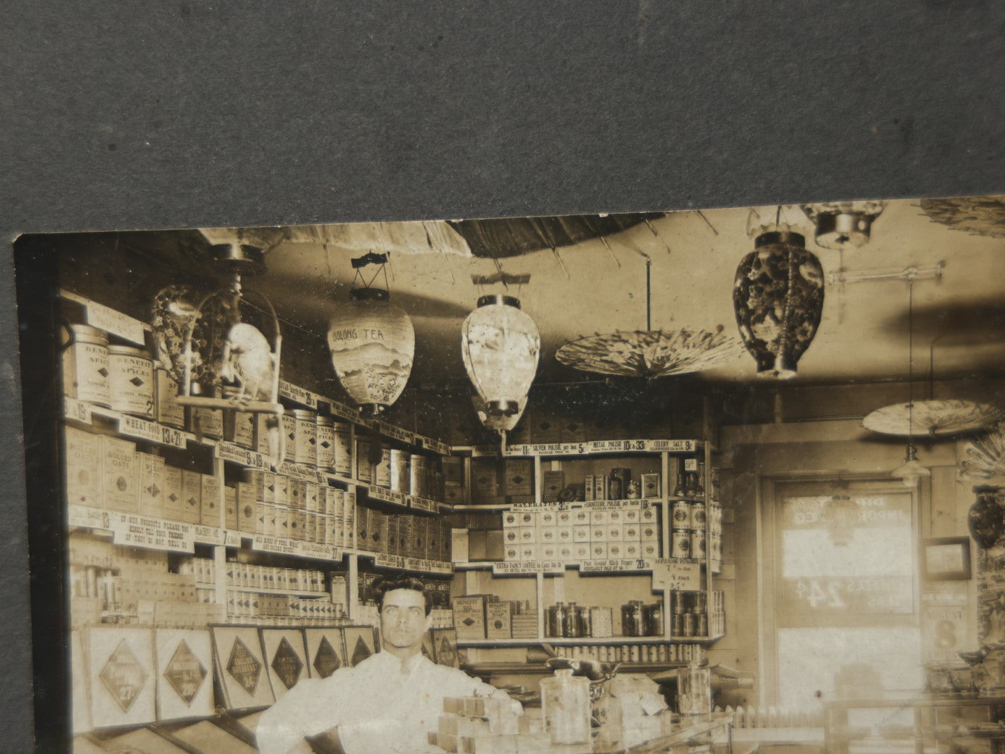 Lot 076 - Antique Boarded Occupational Photograph Of A Handsome Shopkeeper In A General Store, With Advertising, Chinese Lanterns With Tea Ads, And More, Note Bend In Corner