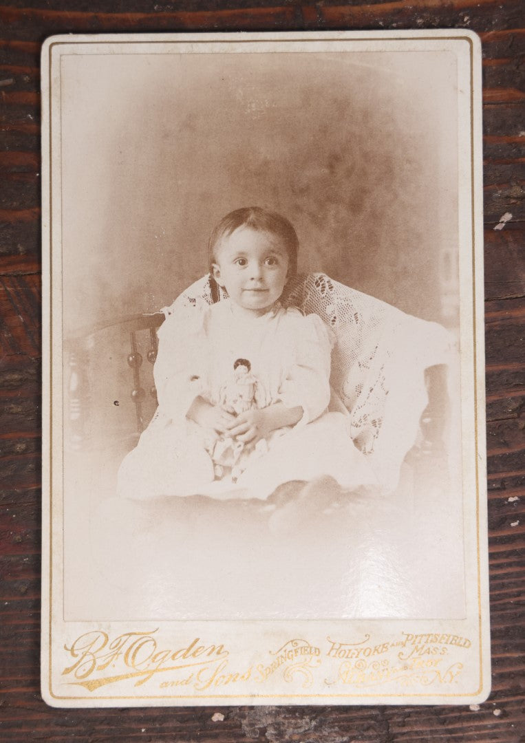 Lot 055 - Antique Cabinet Card Photograph Of A Toddler, Young Boy Or Girl, Holding Doll, With Nervous Smile, Wide Eyes, L.P. Ogden, Successor To B.F. Ogden Photographer, Massachusetts, New York