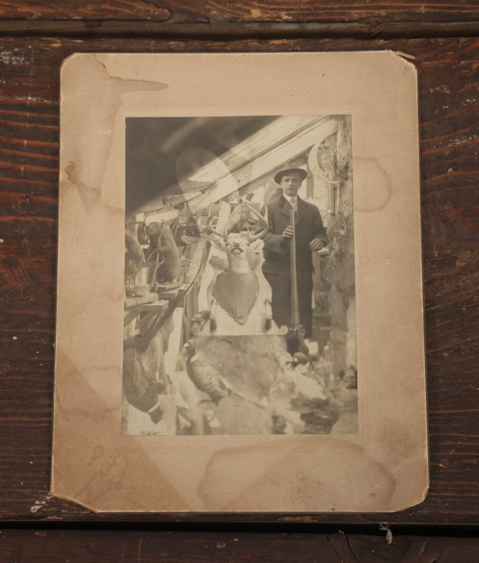 Lot 014 - Antique Boarded Photo Of A Hunter With A Rifle In A Taxidermy Shot, Posing With Buck, Other Mounts Visible Including Beaver, Owl, Etc