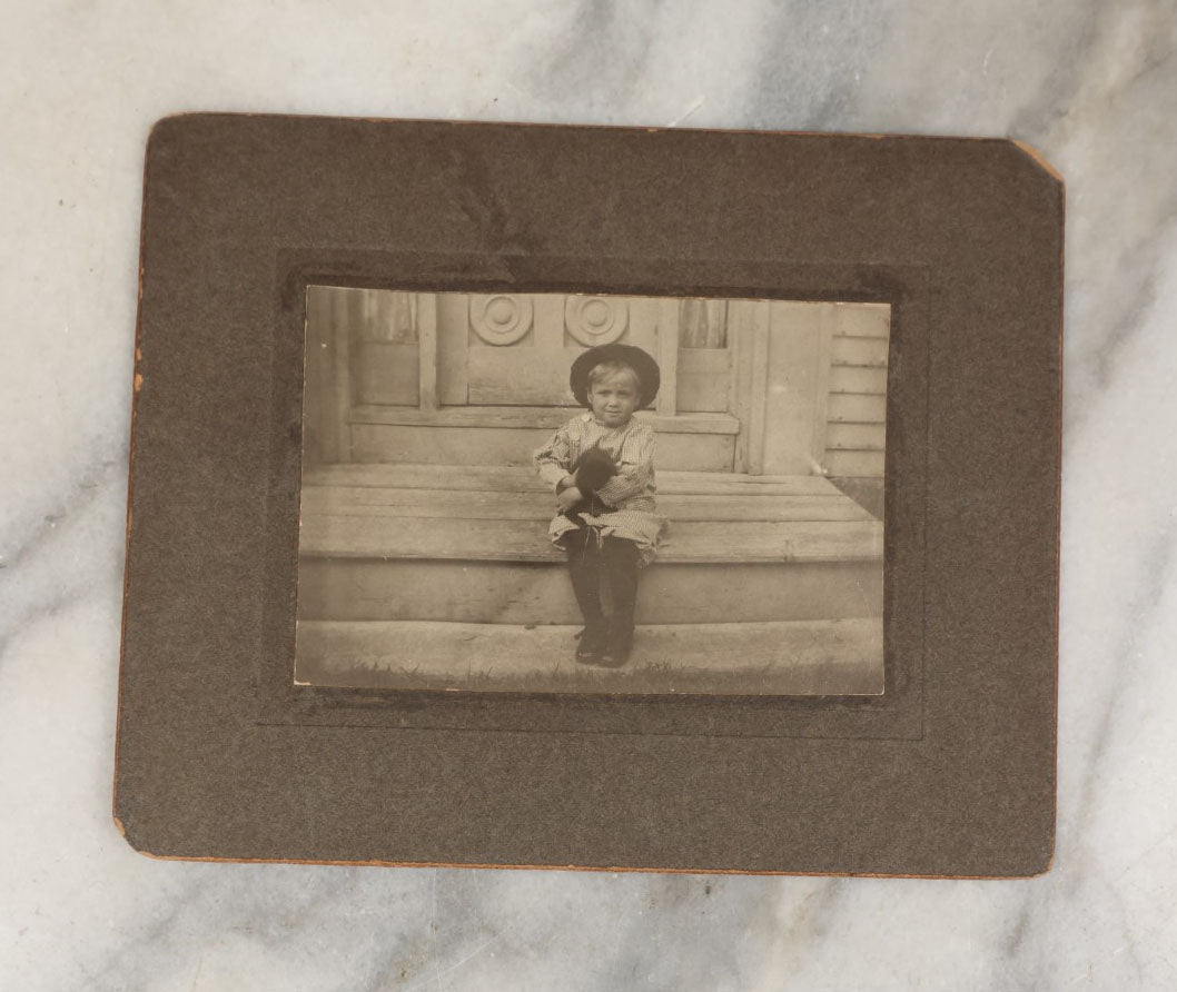 Lot 018 - Antique Boarded Photograph Of A Young Boy In A Hat Sitting On The Front Porch Stoop With A Small Black Cat In His Arms, Cat Moving, Blurry