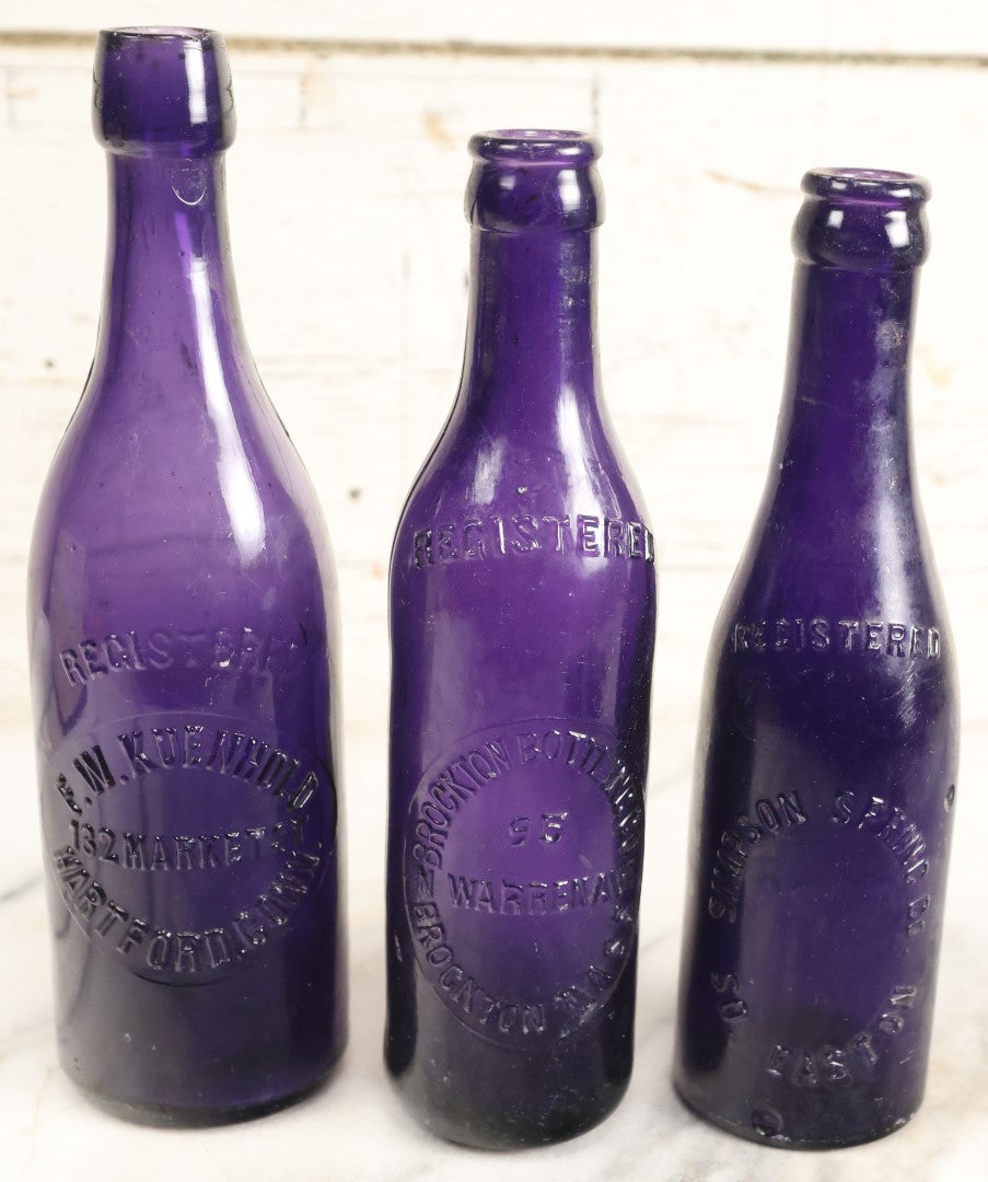 Lot 196 - Grouping Of Three Vintage And Antique Purple Glass Bottles, Including F.W. Kuenhold, Hartford, Connecticut, Sampson Springs Co., South Boston, And Brockton Bottling Co, Massachusetts