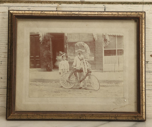 Lot 045 - Antique Framed Photo Of A Young Boy And Man, Likely His Father, With A Bike Covered In Flowers, Flower Moon, Boy In Naval Outfit, Likely For Parade, With Ice Cream Shop In Background