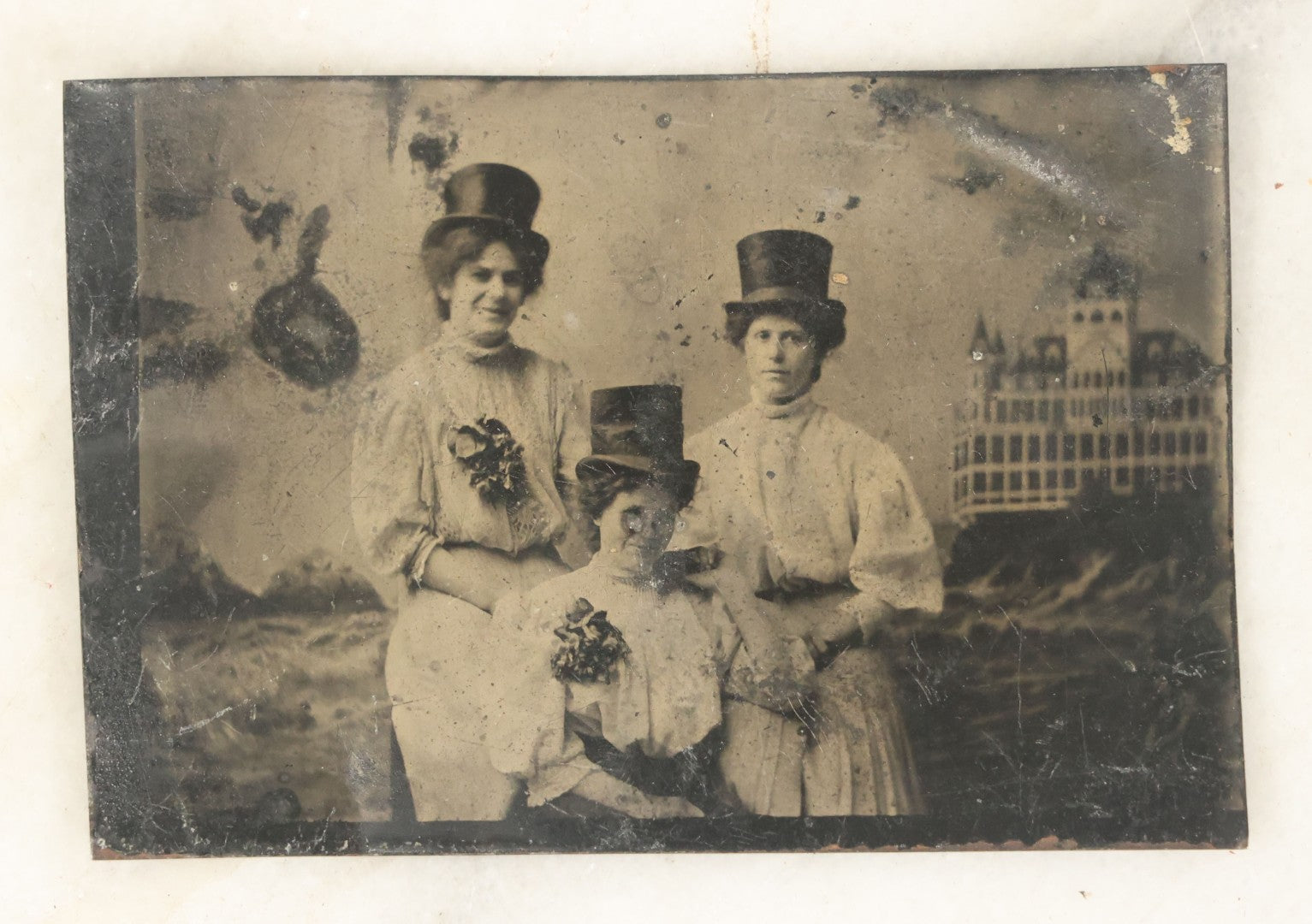 Lot 057 - Single Antique Tintype Photograph, Three Women In Top Hats Posed In Front Of Building Backdrop, With Flowers, Holding Hands