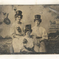 Lot 057 - Single Antique Tintype Photograph, Three Women In Top Hats Posed In Front Of Building Backdrop, With Flowers, Holding Hands