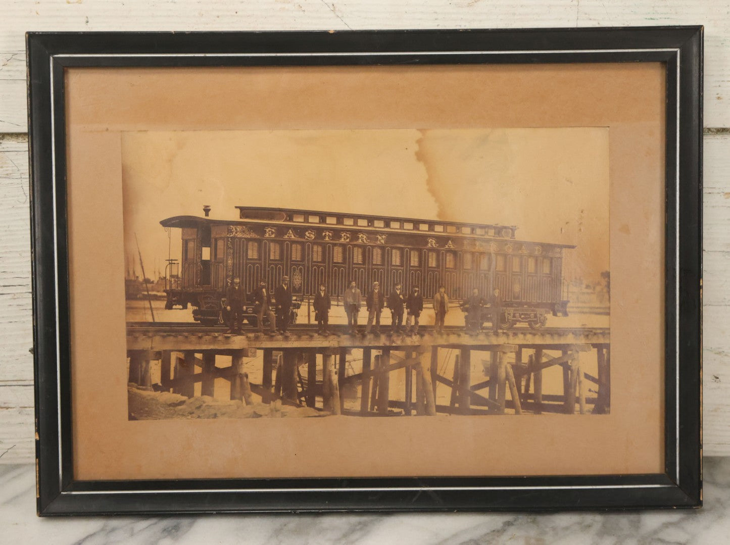 Lot 037 - Antique Boarded Occupational Photograph Of Newly Built Eastern Railroad Passenger Train Car, With Workers Pictured On Train Track, In Front Of Harbor, In Frame, Note Water Stains, 15-5/8" x 11-1/4"