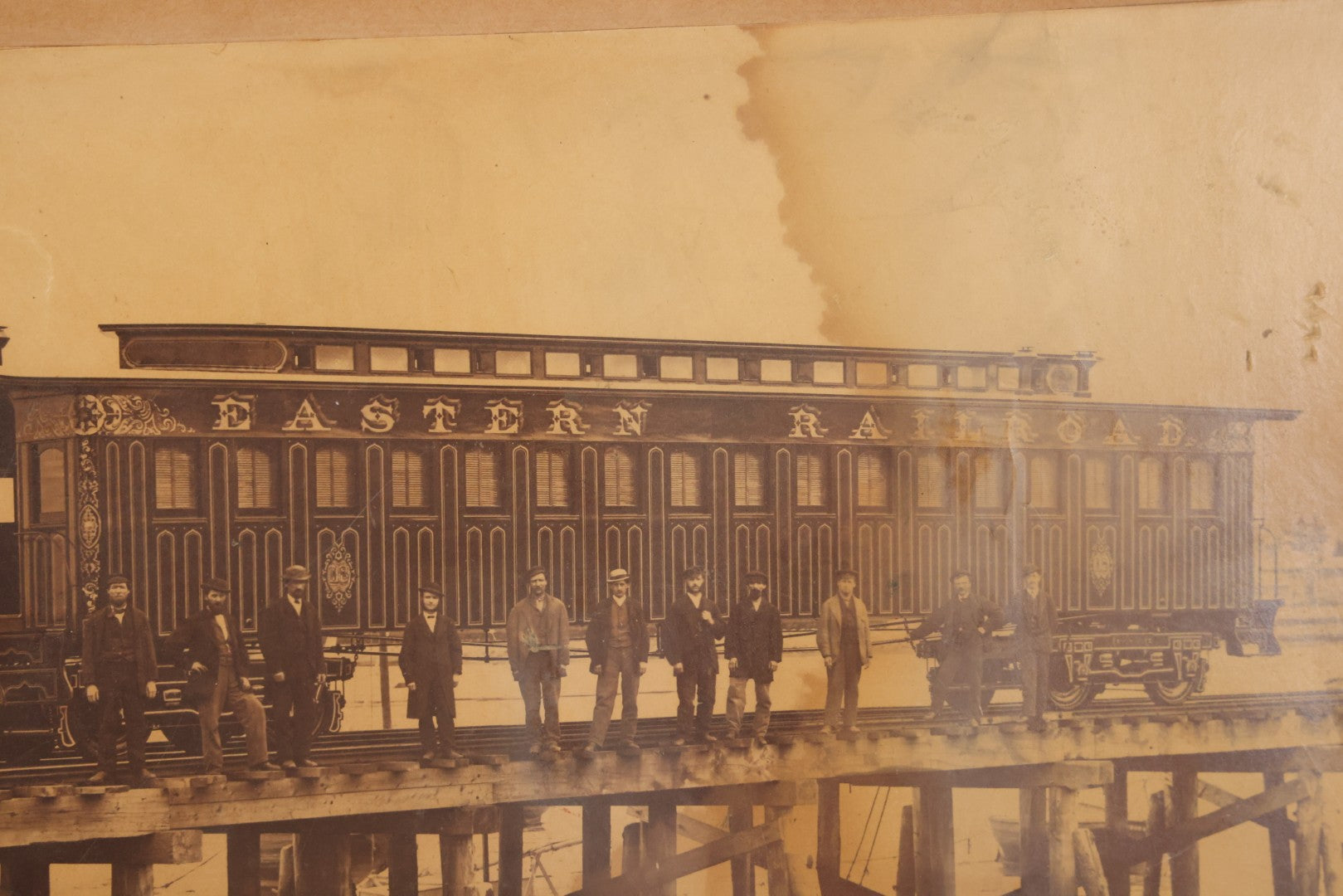 Lot 037 - Antique Boarded Occupational Photograph Of Newly Built Eastern Railroad Passenger Train Car, With Workers Pictured On Train Track, In Front Of Harbor, In Frame, Note Water Stains, 15-5/8" x 11-1/4"