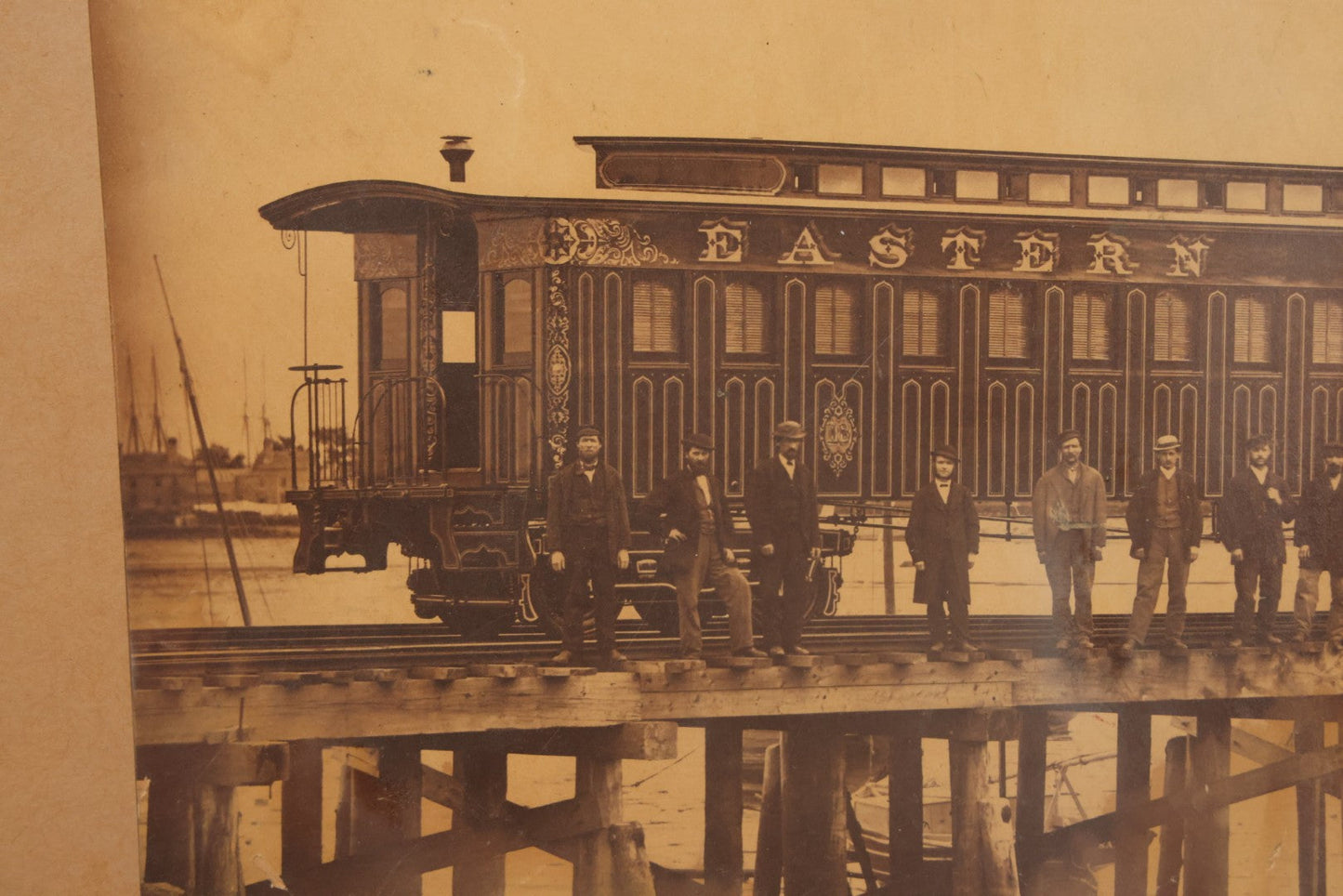 Lot 037 - Antique Boarded Occupational Photograph Of Newly Built Eastern Railroad Passenger Train Car, With Workers Pictured On Train Track, In Front Of Harbor, In Frame, Note Water Stains, 15-5/8" x 11-1/4"