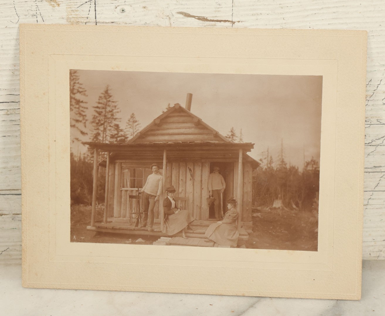 Lot 096 - Antique Boarded Photograph Of Two Men And Two Women Posing On The Stoop Of A Hunting Cabin, Men Holding Rifles