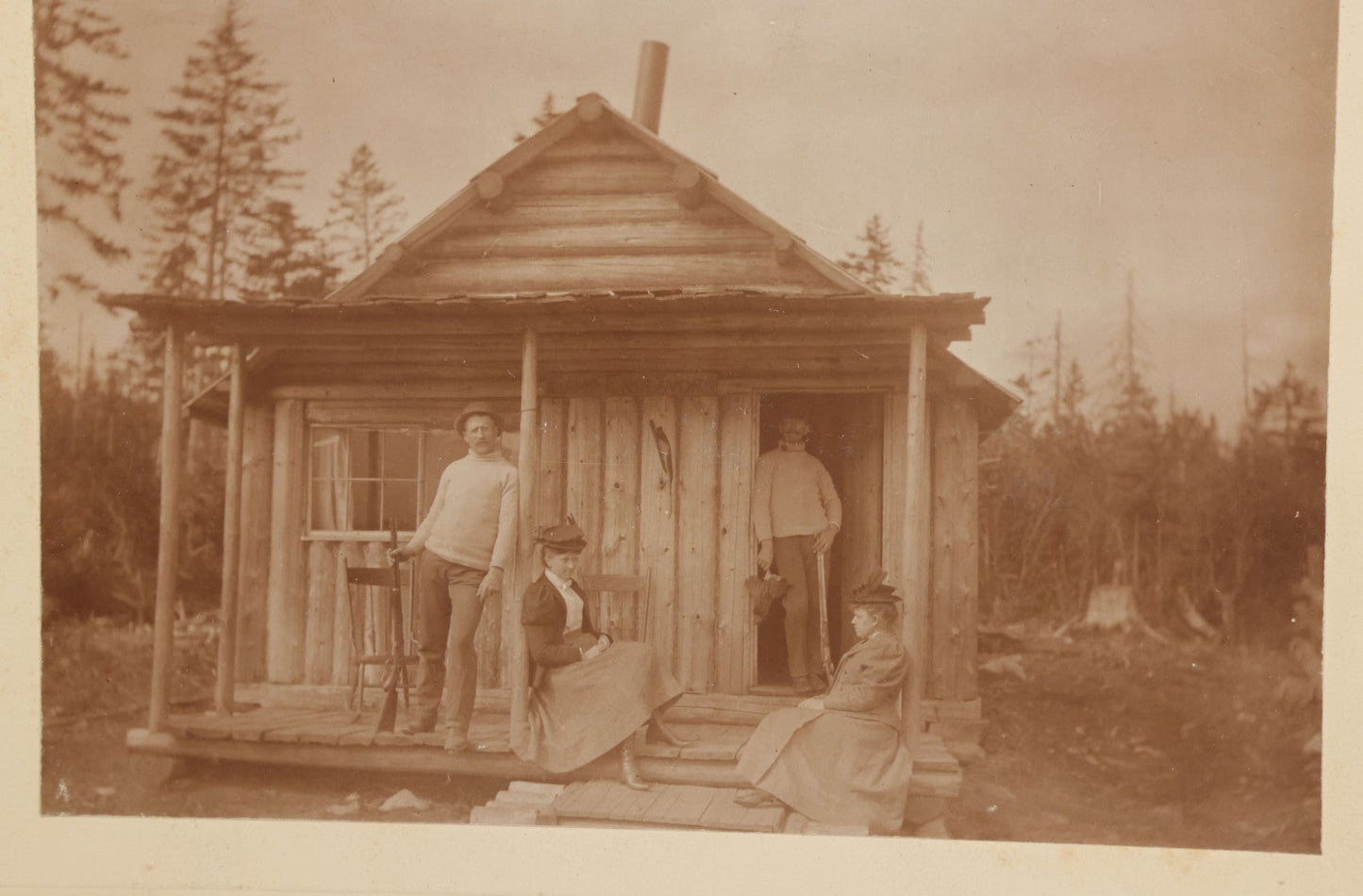 Lot 096 - Antique Boarded Photograph Of Two Men And Two Women Posing On The Stoop Of A Hunting Cabin, Men Holding Rifles