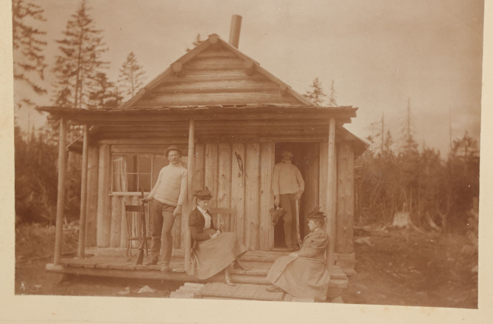 Lot 096 - Antique Boarded Photograph Of Two Men And Two Women Posing On The Stoop Of A Hunting Cabin, Men Holding Rifles