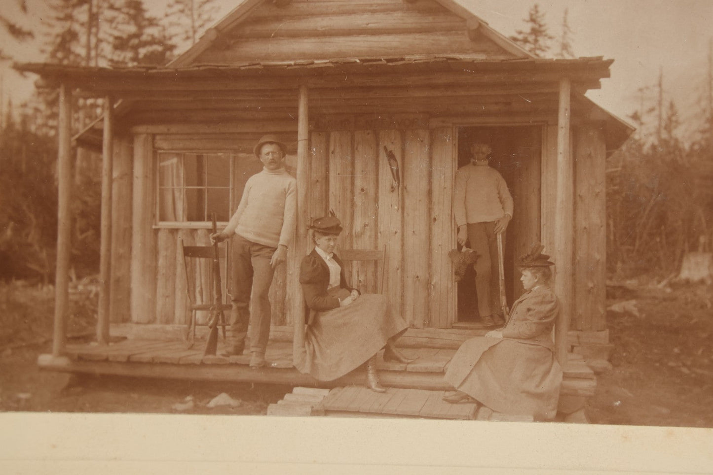 Lot 096 - Antique Boarded Photograph Of Two Men And Two Women Posing On The Stoop Of A Hunting Cabin, Men Holding Rifles