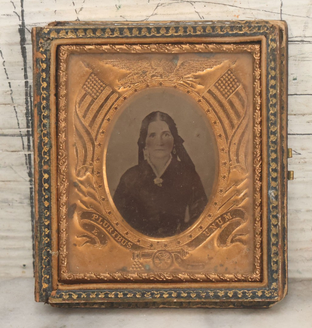 Lot 117 - Antique 1/6th Plate Ambrotype Photograph Of Solemn Woman In Dark Clothing With Patriotic Civil War Era Brass Mat With American Flags, Eagle, Cannon, "E Pluribus Unum" Banner, In Half Case