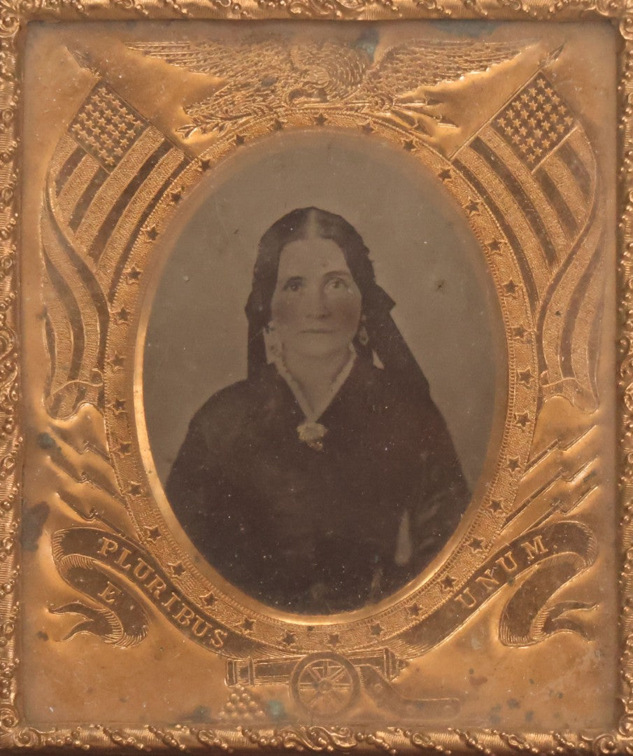 Lot 117 - Antique 1/6th Plate Ambrotype Photograph Of Solemn Woman In Dark Clothing With Patriotic Civil War Era Brass Mat With American Flags, Eagle, Cannon, "E Pluribus Unum" Banner, In Half Case