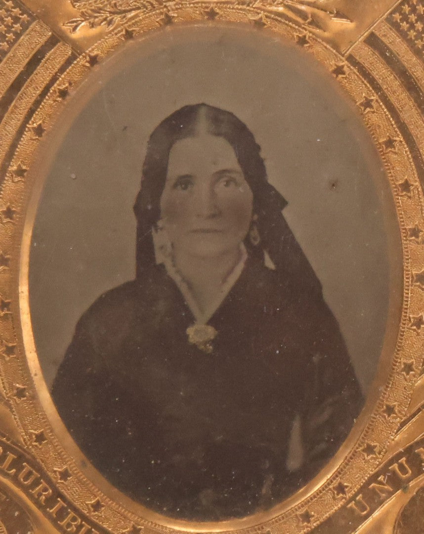 Lot 117 - Antique 1/6th Plate Ambrotype Photograph Of Solemn Woman In Dark Clothing With Patriotic Civil War Era Brass Mat With American Flags, Eagle, Cannon, "E Pluribus Unum" Banner, In Half Case