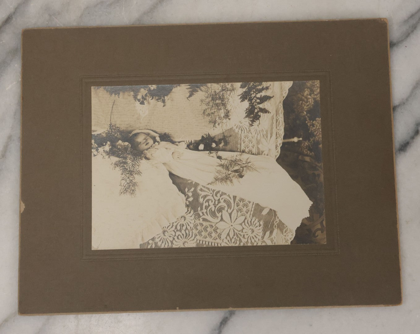 Lot 030 - Antique Boarded Post Mortem Photograph Of Baby In Long White Robes, Laid Out On Lace Floral Linens With Flowers