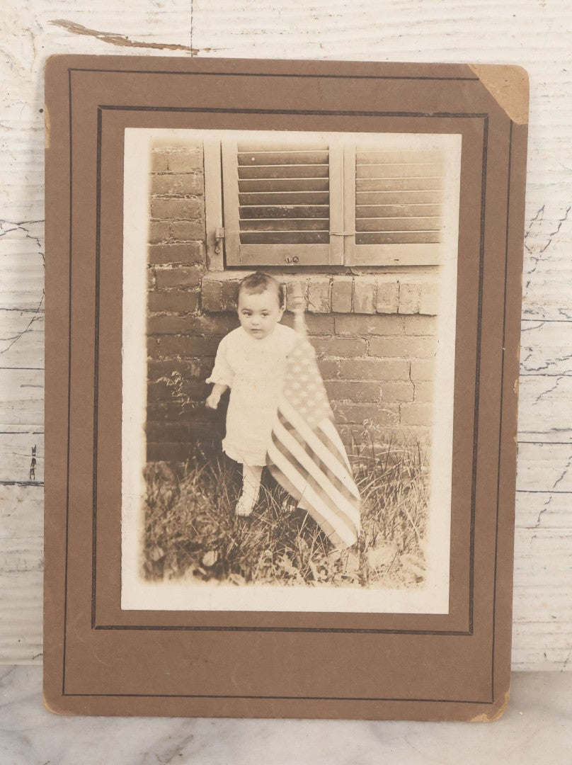 Lot 075 - Antique Boarded Photograph Of Toddler Standing Behind An American Flag In Front Of A Brick Wall With Shutters