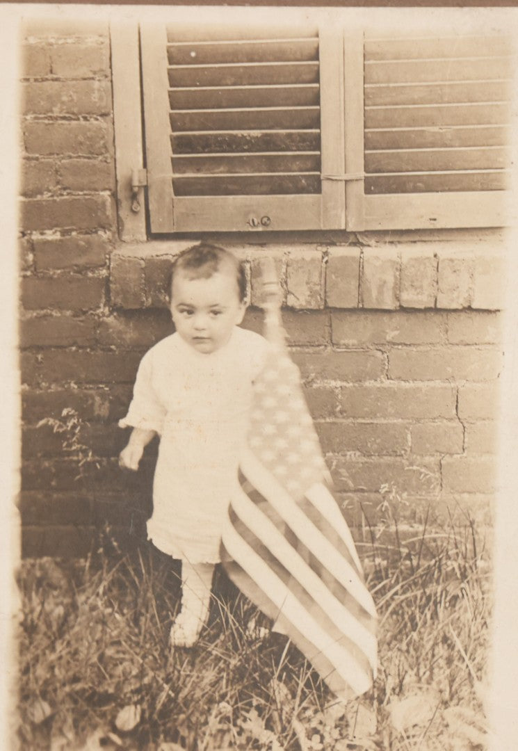 Lot 075 - Antique Boarded Photograph Of Toddler Standing Behind An American Flag In Front Of A Brick Wall With Shutters