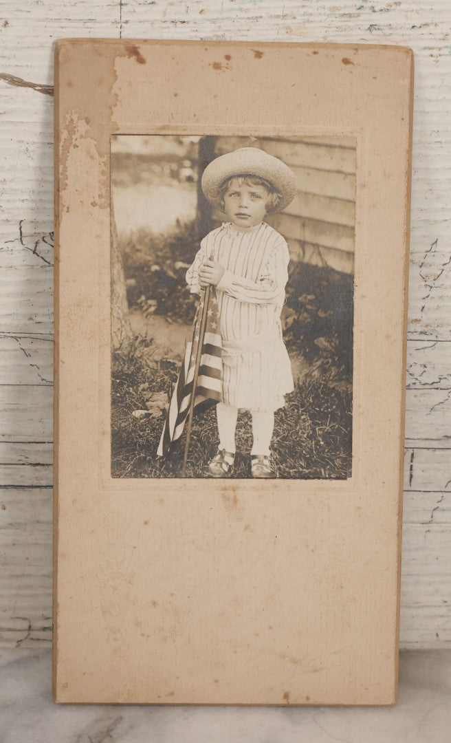 Lot 076 - Antique Boarded Photograph Of Young Child In Striped Shirt And Hat Standing With An American Flag In The Grass
