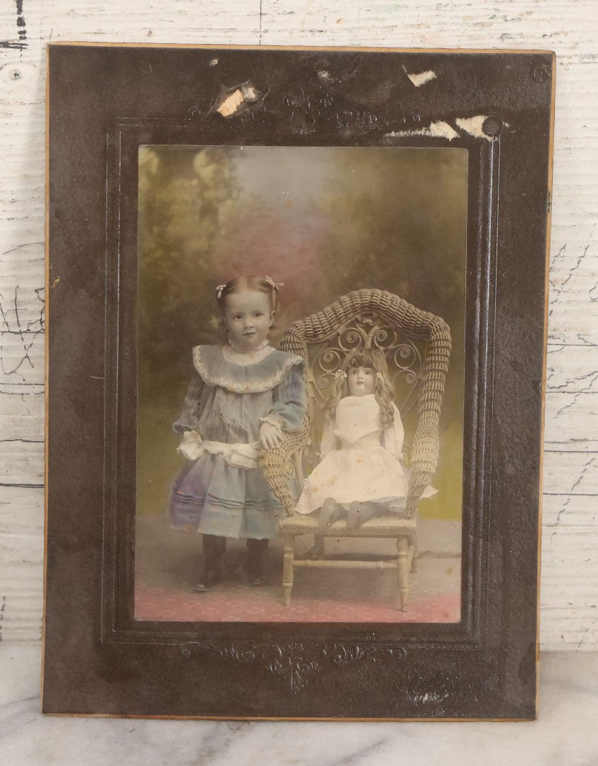 Lot 077 - Antique Boarded Tinted Photograph Of Young Girl In Blue Dress Standing Besides Wicker Chair With Her Period China Doll, Note Color Tinting Notes On Verso