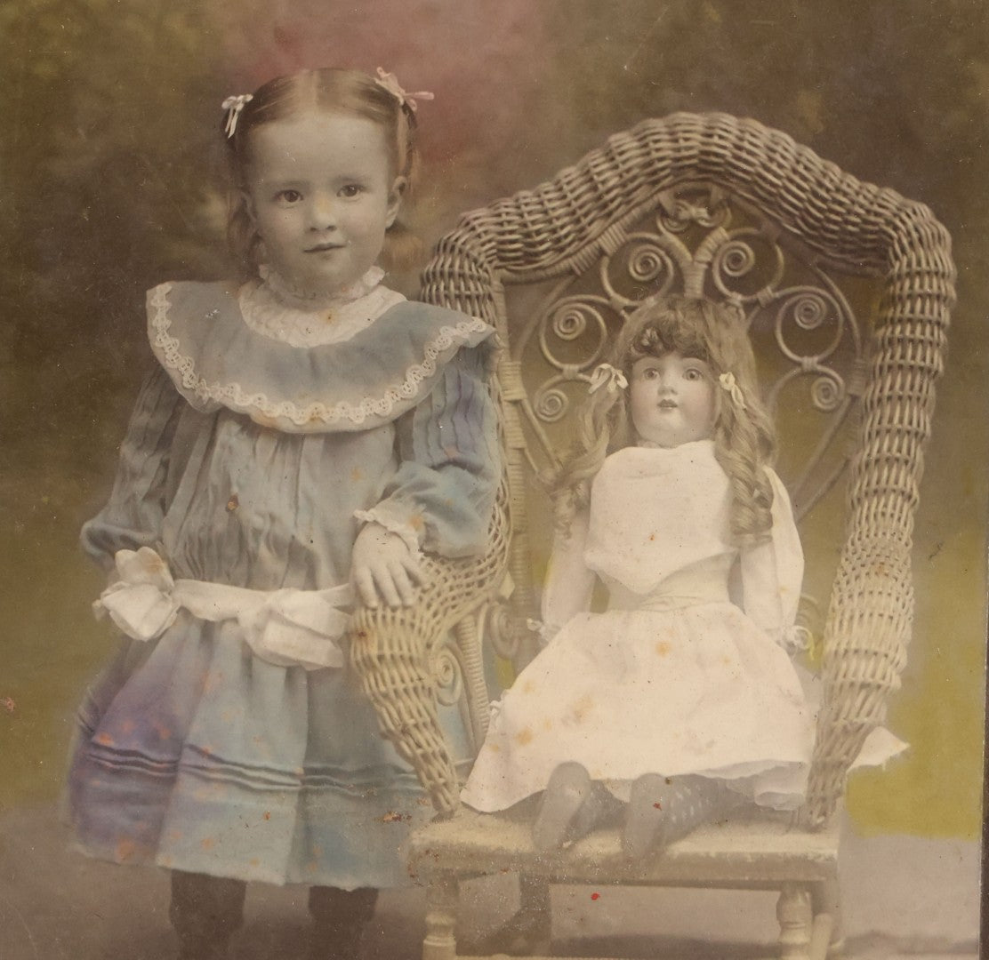 Lot 077 - Antique Boarded Tinted Photograph Of Young Girl In Blue Dress Standing Besides Wicker Chair With Her Period China Doll, Note Color Tinting Notes On Verso