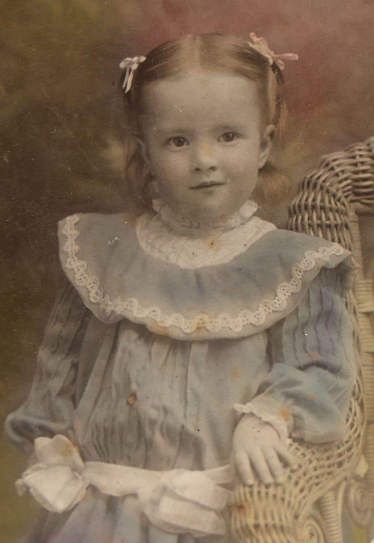 Lot 077 - Antique Boarded Tinted Photograph Of Young Girl In Blue Dress Standing Besides Wicker Chair With Her Period China Doll, Note Color Tinting Notes On Verso