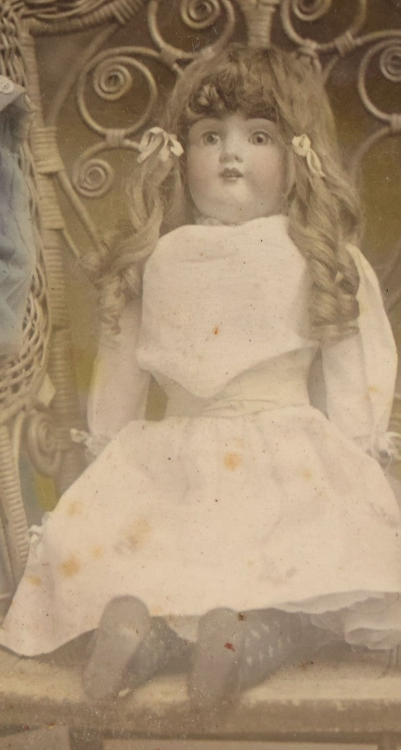 Lot 077 - Antique Boarded Tinted Photograph Of Young Girl In Blue Dress Standing Besides Wicker Chair With Her Period China Doll, Note Color Tinting Notes On Verso