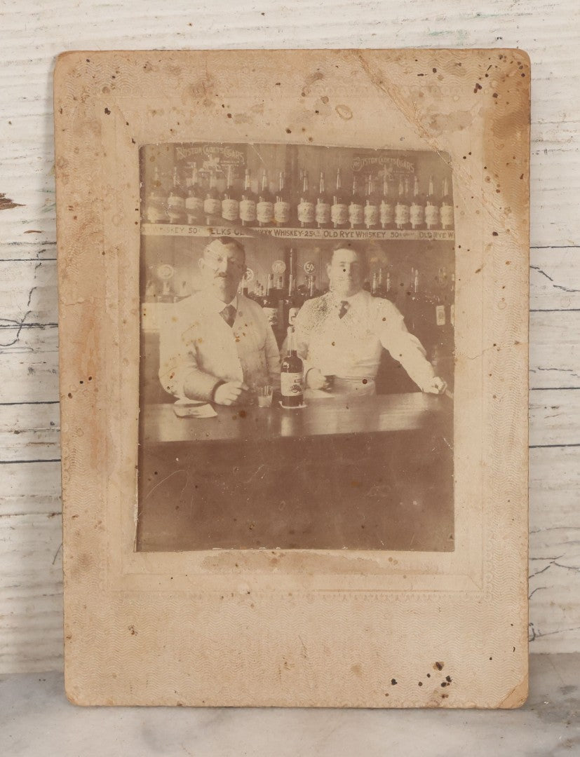 Lot 083 - Antique Boarded Occupational Photograph Of Two Bartenders Standing Behind Bar Posing With Bottles Of Elks Club Whiskey, Other Advertisements Visible Including For Boston Cadet Cigars, Note Damaged Upper Right Corner