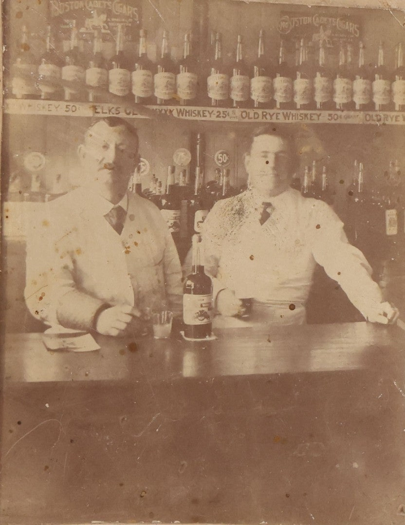 Lot 083 - Antique Boarded Occupational Photograph Of Two Bartenders Standing Behind Bar Posing With Bottles Of Elks Club Whiskey, Other Advertisements Visible Including For Boston Cadet Cigars, Note Damaged Upper Right Corner
