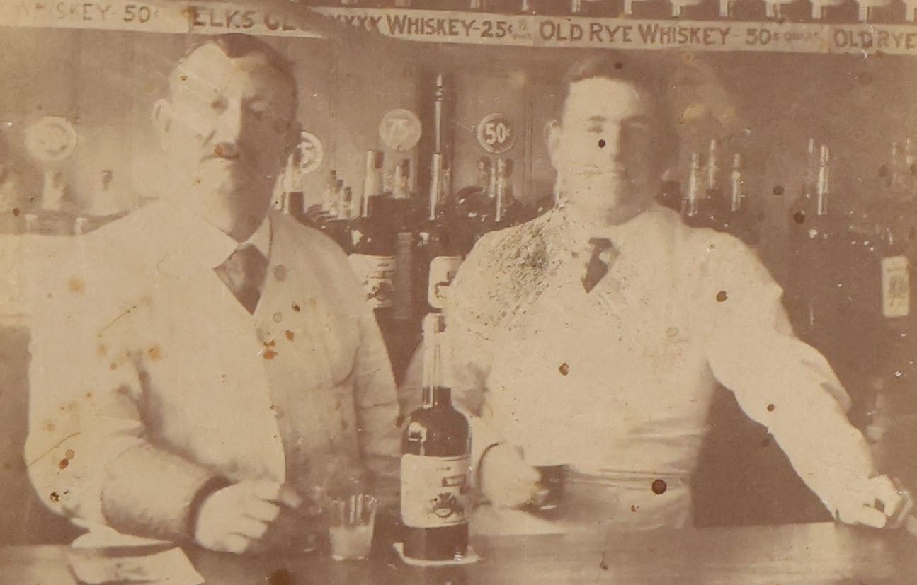 Lot 083 - Antique Boarded Occupational Photograph Of Two Bartenders Standing Behind Bar Posing With Bottles Of Elks Club Whiskey, Other Advertisements Visible Including For Boston Cadet Cigars, Note Damaged Upper Right Corner