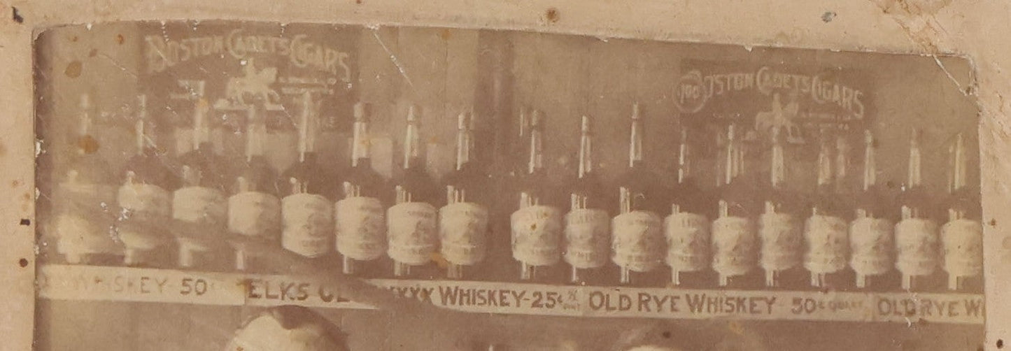 Lot 083 - Antique Boarded Occupational Photograph Of Two Bartenders Standing Behind Bar Posing With Bottles Of Elks Club Whiskey, Other Advertisements Visible Including For Boston Cadet Cigars, Note Damaged Upper Right Corner
