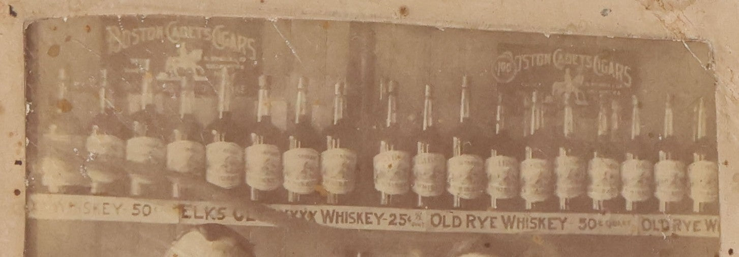 Lot 083 - Antique Boarded Occupational Photograph Of Two Bartenders Standing Behind Bar Posing With Bottles Of Elks Club Whiskey, Other Advertisements Visible Including For Boston Cadet Cigars, Note Damaged Upper Right Corner