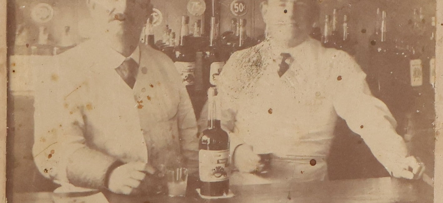 Lot 083 - Antique Boarded Occupational Photograph Of Two Bartenders Standing Behind Bar Posing With Bottles Of Elks Club Whiskey, Other Advertisements Visible Including For Boston Cadet Cigars, Note Damaged Upper Right Corner