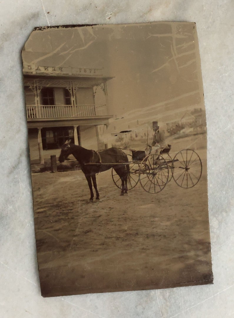 Lot 074 - Antique Outdoor Tintype Photograph Of Man In Stovepipe Hat Sitting On Horse Drawn Cart In Front Of A Building With Partial Sign Visible And Houses In Background