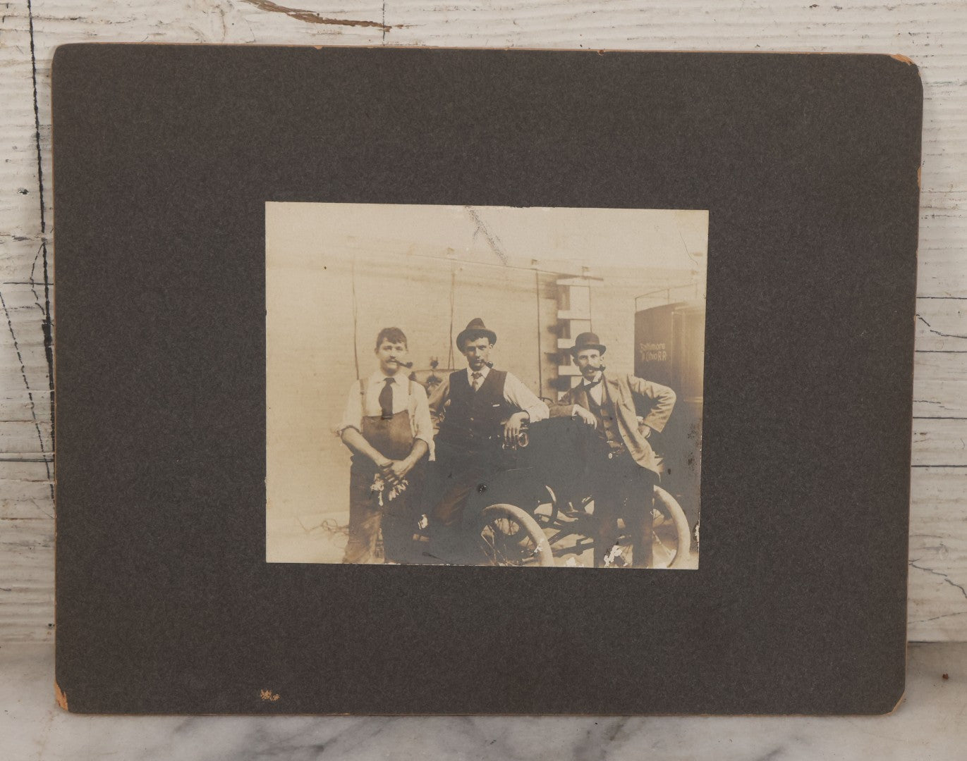 Lot 072 - Antique Boarded Occupational Photograph Of Three Men Smoking Pipes And Posing With Turn Of The Century Automobile, With Baltimore & Ohio Railroad Carriage Visible In Background, Handwritten Note On Verso, Dated October 9, 1901