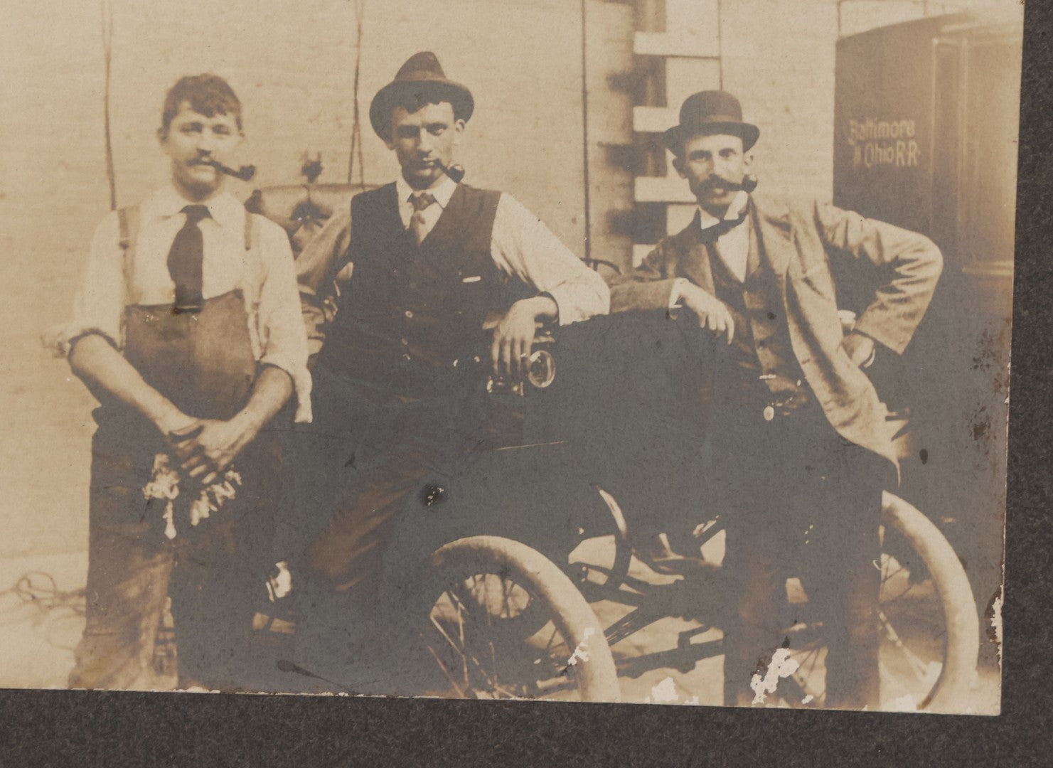 Lot 072 - Antique Boarded Occupational Photograph Of Three Men Smoking Pipes And Posing With Turn Of The Century Automobile, With Baltimore & Ohio Railroad Carriage Visible In Background, Handwritten Note On Verso, Dated October 9, 1901