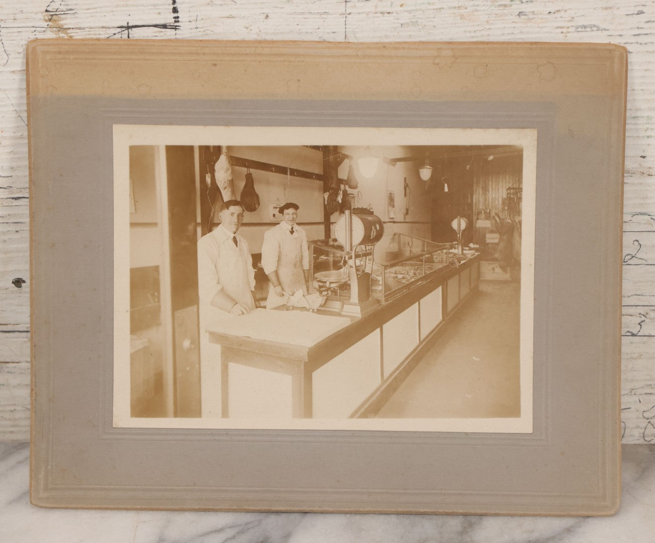 Lot 077 - Antique Boarded Occupational Photograph Of Two Butchers Behind Counter, With Sausage, Full Pig, And Other Meat Products Visible In Photo, And Large Commercial Scale