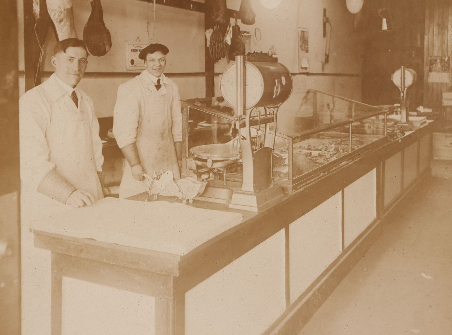 Lot 077 - Antique Boarded Occupational Photograph Of Two Butchers Behind Counter, With Sausage, Full Pig, And Other Meat Products Visible In Photo, And Large Commercial Scale