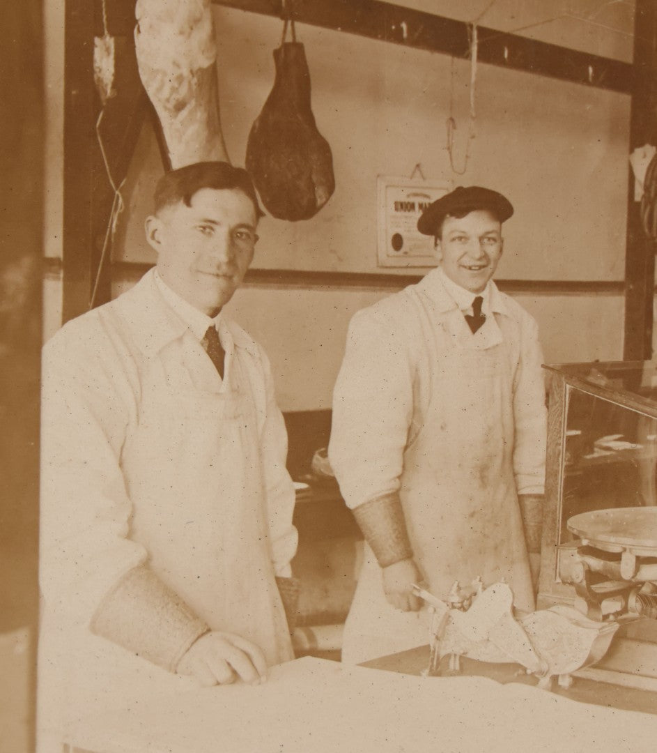 Lot 077 - Antique Boarded Occupational Photograph Of Two Butchers Behind Counter, With Sausage, Full Pig, And Other Meat Products Visible In Photo, And Large Commercial Scale