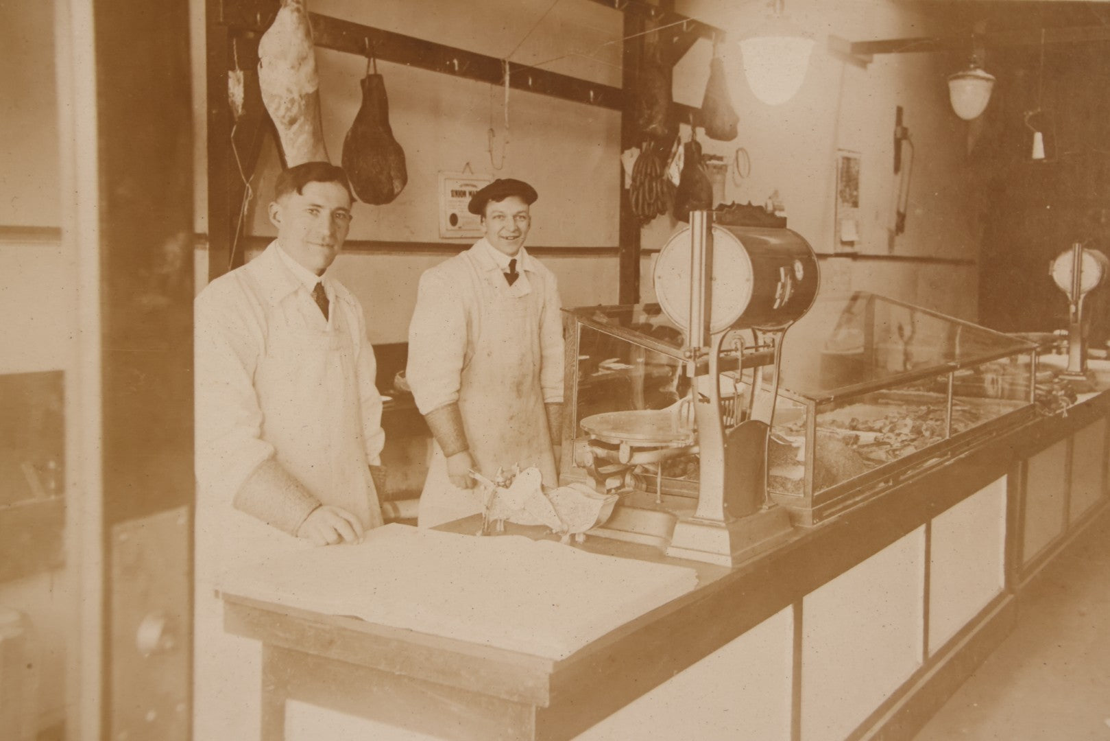 Lot 077 - Antique Boarded Occupational Photograph Of Two Butchers Behind Counter, With Sausage, Full Pig, And Other Meat Products Visible In Photo, And Large Commercial Scale