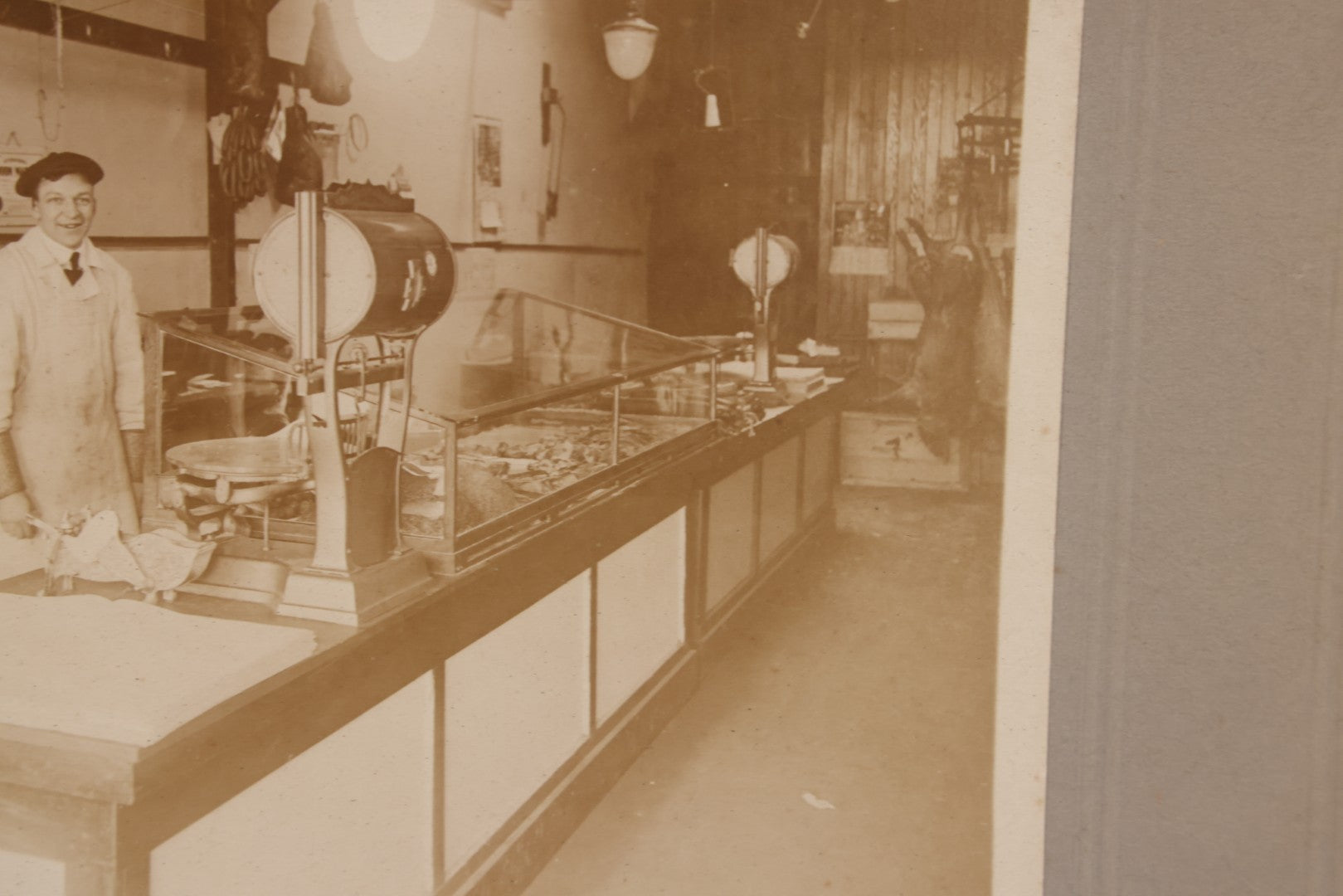 Lot 077 - Antique Boarded Occupational Photograph Of Two Butchers Behind Counter, With Sausage, Full Pig, And Other Meat Products Visible In Photo, And Large Commercial Scale