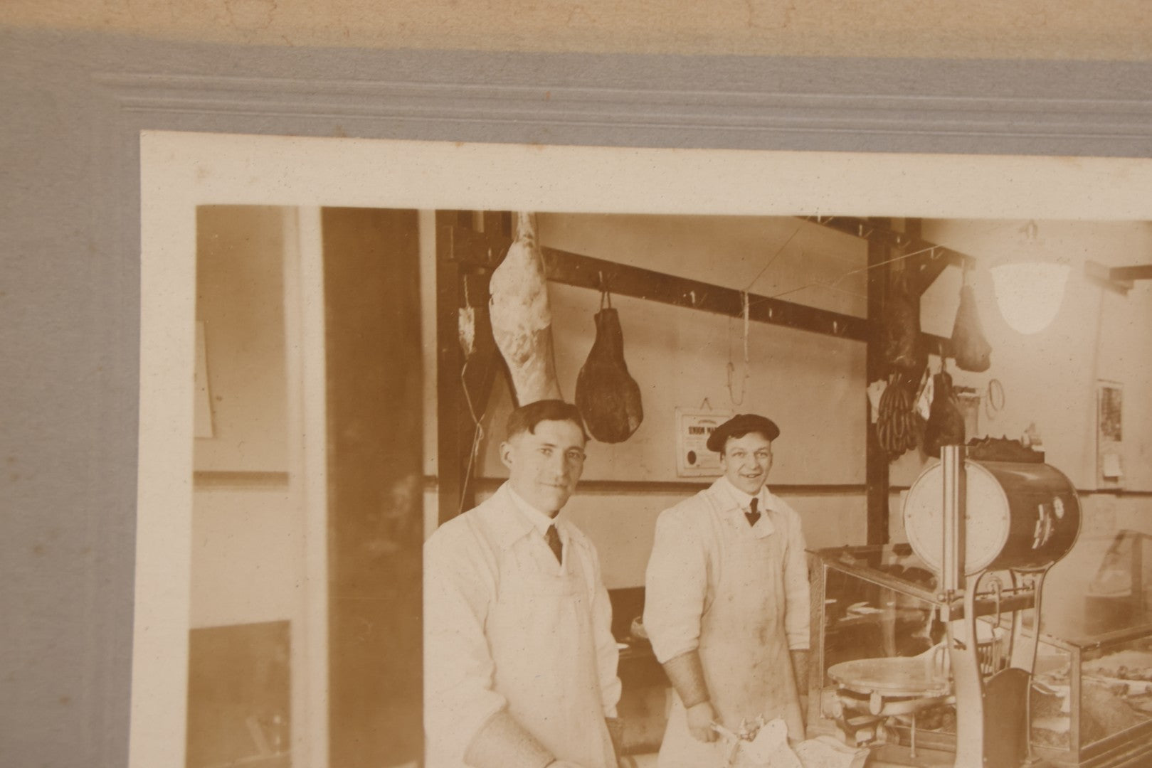 Lot 077 - Antique Boarded Occupational Photograph Of Two Butchers Behind Counter, With Sausage, Full Pig, And Other Meat Products Visible In Photo, And Large Commercial Scale