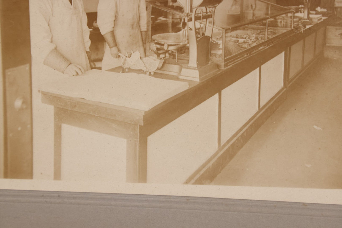 Lot 077 - Antique Boarded Occupational Photograph Of Two Butchers Behind Counter, With Sausage, Full Pig, And Other Meat Products Visible In Photo, And Large Commercial Scale
