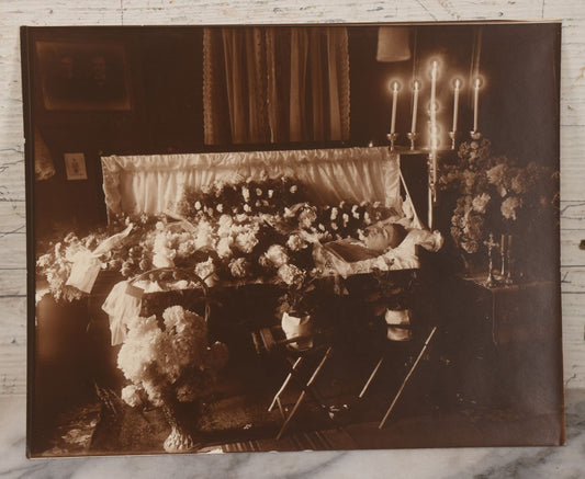 Lot 101 - Vintage Paper Funeral Photograph Of Young Man In Coffin Adorned With Floral Arrangements And "Dear Brother" Ribbon, Crosses, Photos, Candles And Religious Art Visible, Including Photo Of WWI Soldier In Top Left