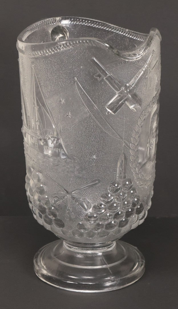 Lot 027 - Antique Early American Pattern Glass E.A.P.G. Spanish American War Battle Of Manila Bay Commemorative Pitcher With Admiral Dewey And U.S.S. Olympia, Patriotic Symbols Including Weaponry And Flags, Manufactured By Beatty-Brady Glass Co., Indiana