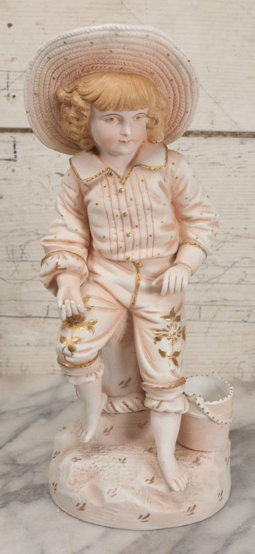 Lot 037 - Vintage Bisque Statue Of Young Blond Child In Large Straw Hat Sitting By Shore With Pail, With Pink Painted Details And Gold Embellishments, Note Chip, Repairs, 12" Height