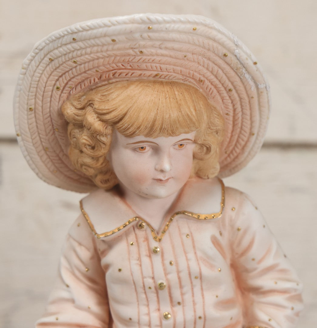 Lot 037 - Vintage Bisque Statue Of Young Blond Child In Large Straw Hat Sitting By Shore With Pail, With Pink Painted Details And Gold Embellishments, Note Chip, Repairs, 12" Height